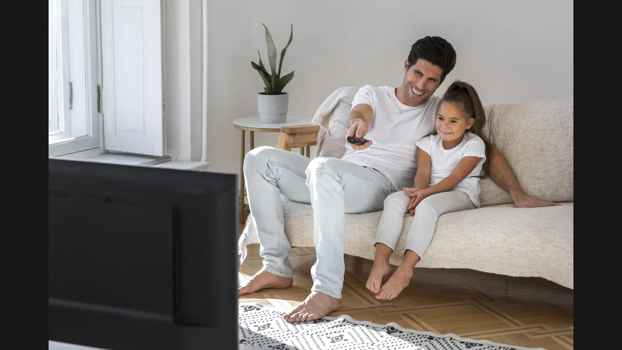 A father and his young daughter sitting on a sofa watching television, the father holding a remote