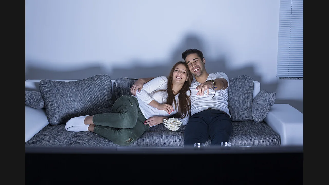A young couple with a bowl of popcorn watching television on a sofa at night